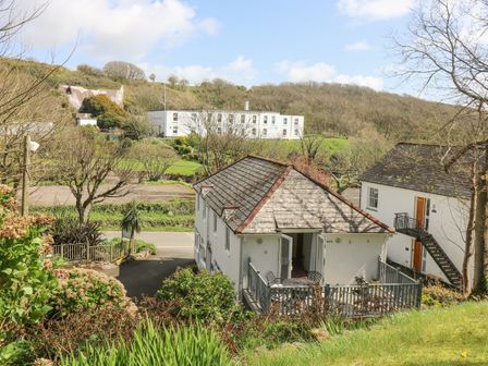 A house with a patio and outdoor furniture in a rural area with trees and a large white building in the background at Longships in Porthcurno