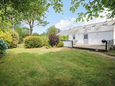 A garden with grass trees bushes and a white house with a deck and outdoor furniture at Little Chygowlin in Truro