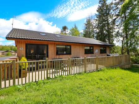 A wooden cabin with a fenced porch and a grassy yard surrounded by trees at Broken-Sky Lodge in Otterburn