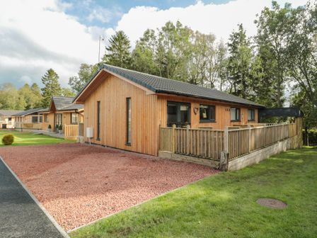 A single-story wooden house with a fenced porch and gravel driveway surrounded by grass and trees at Barn Owl in Otterburn