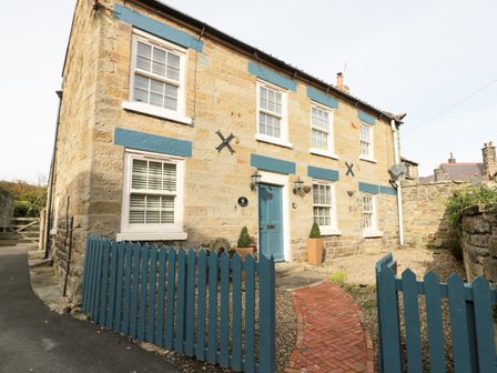 A stone house with blue window trims and a blue front door with a matching blue picket fence and brick path at The Nook at 29 High Street in Cloughton near Burniston