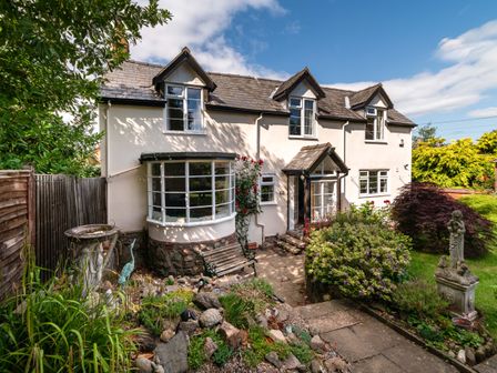 A garden with plants and statues in front of a two story house with dormer windows and a bay window at Eleri Cottage in Great Malvern