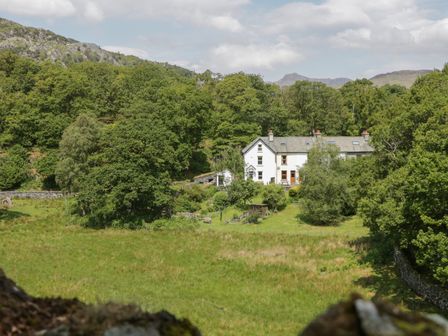 A white house surrounded by trees and grass with hills in the background at Acorn Cottage in Coniston