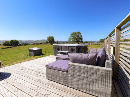 An outdoor wooden deck with a wicker sofa and a hot tub overlooking a grassy field at The Wren in Llysfaen