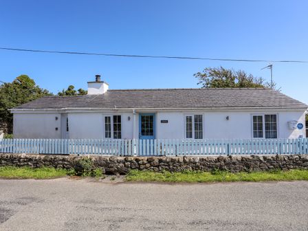A front view of a single-story house with a blue door white walls and a blue picket fence at Ffrwd in Rhosneigr