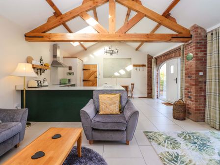 A living area with exposed wooden beams a gray armchair a wooden coffee table and an open kitchen with green cabinets at Plough Cottage in Little Kelk near Bridlington