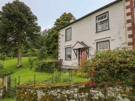 An exterior view of a white house with black window frames a red door and a small porch surrounded by green grass trees and bushes at High Torver House in Torver