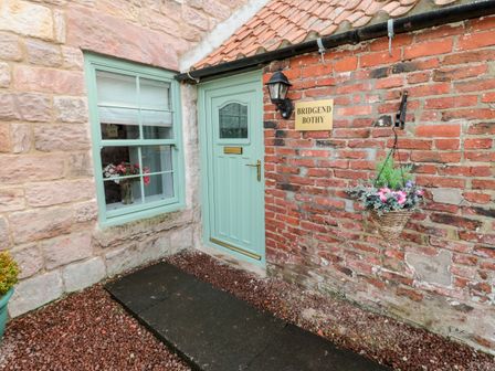 A green door and window on a brick house with a hanging basket and a sign that reads Bridgend Bothy in Wooler