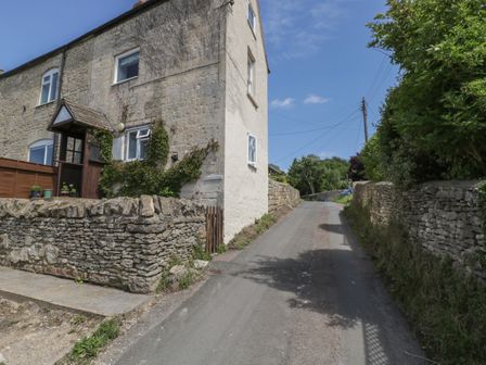 A stone building with plants growing on the wall next to a narrow paved road with stone walls on both sides at Athelstan Cottage Chloe Mead near Stroud