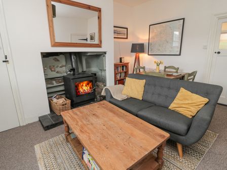 A living room with a wood stove, a grey sofa with yellow cushions, a wooden coffee table, a dining table with chairs, a floor lamp, and framed pictures on the walls at Lavender Cottage in Glanton