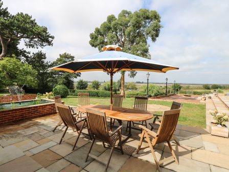 An outdoor patio with a wooden table and chairs under a large umbrella next to a small pond at Waveney View Cottage in Burgh Castle