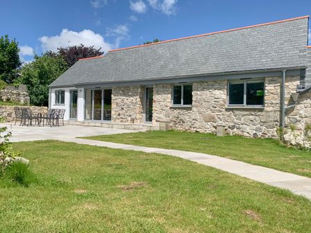 A stone house with a slate roof and patio furniture on a tiled patio with a lawn and walkway at The Corn Store in St Stephen
