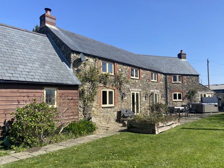 An exterior view of a stone house with a slate roof a garden patio with chairs and a hot tub at Varley Lodge in Prixford near Barnstaple