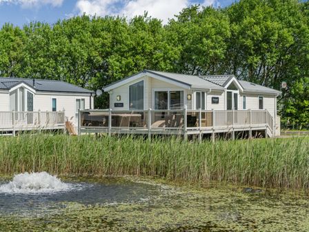 Two white lodges with decks near a pond with plants and trees in the background at Links Lodge in Flookburgh