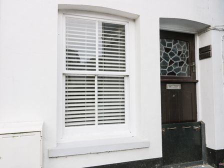 A white wall with a window featuring horizontal blinds and a dark wooden door with decorative glass at Bay Tree Cottage in West Looe