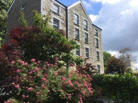 A stone building with multiple windows surrounded by green and pink flowering bushes at The Old Mill in Neyland near Milford Haven