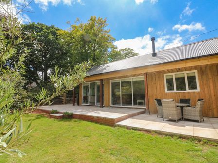 A wooden house with sliding glass doors a patio with outdoor seating and a garden at Langdale Barn in Sampford Courtenay