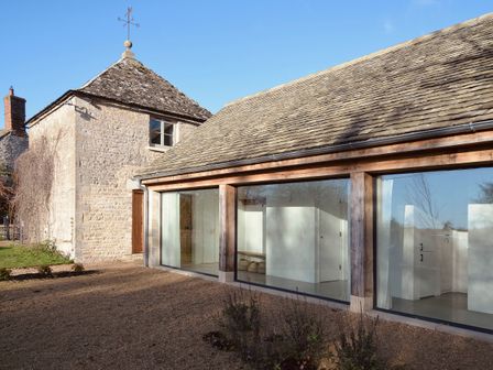 A stone building with a weather vane and a modern extension with large glass windows at High Cogges Farm Holiday Cottages - The Cart Shed in High Cogges near Witney