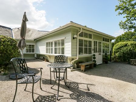 An outdoor patio area with a metal table and chairs and a closed umbrella outside a house with white siding at Woodland Lodge in Meathop near Grange-Over-Sands
