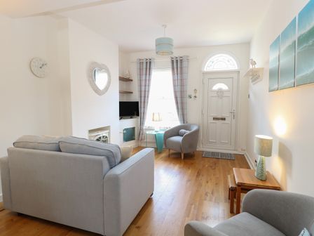 A living room with two grey armchairs a grey sofa a white door with a window and curtains at Gull's Nest in Pakefield