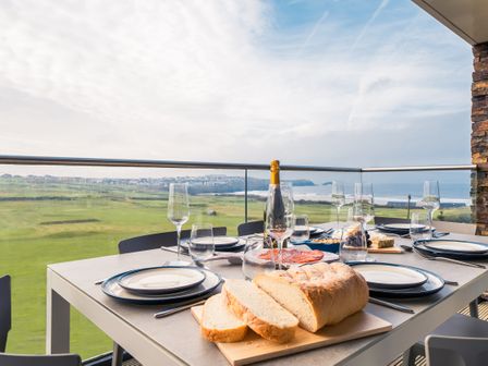 A dining table with plates glasses bread and a bottle of wine on a balcony overlooking green fields and the sea at Newquay Fistral Beach View in Newquay