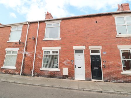 A row of brick terraced houses with white and black front doors and windows on a paved street at Sandy Toes Cottage in Newbiggin-By-The-Sea