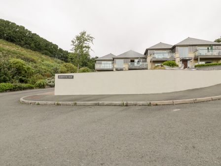 Residential buildings with balconies behind a white wall with a morweth view sign and a sloped road with greenery on one side at The Bolt Hole in Downderry