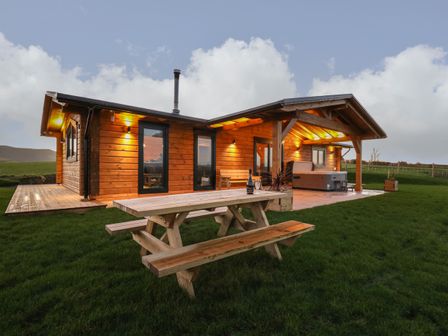 An outdoor area with a wooden picnic table a wooden cabin with exterior lights and a covered patio with a hot tub at Bacheiddon Log Cabin in Aberhosan near Machynlleth