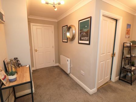 A hallway with two white doors a small wooden table with decorations a black shelving unit with plants framed pictures and a round mirror on the wall at Esk Retreat in Whitby