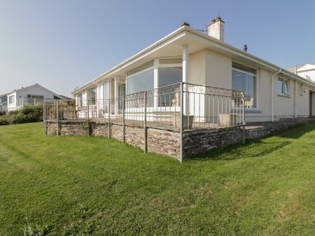 An exterior view of a single-story house with a stone foundation and metal railing on a grassy lawn at Gwel An Mor in Newquay