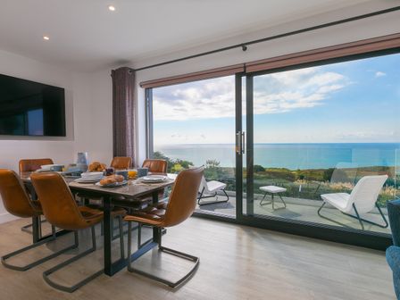 A dining area with table and chairs set for breakfast next to large sliding glass doors overlooking the sea at Sea Sands in Praa Sands