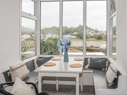A dining nook with a white table and wicker placemats surrounded by black bench seating and cushions with a vase of blue flowers at Ivy Cottage in Borth-y-Gest near Porthmadog
