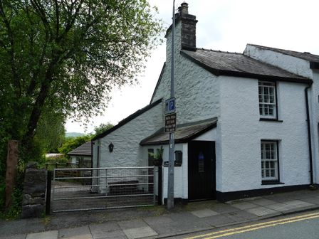 A white house with a black door and windows next to a tree and a metal gate by the roadside at Glan Y Ddol in Trefriw