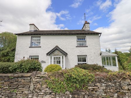 A white two-story house with chimneys and a small porch behind a stone wall with greenery at Greenstyles in Bowness-On-Windermere