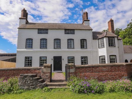 A white two-story house with multiple windows and chimneys behind a brick wall with flowers at Plas Coch Near St Asaph