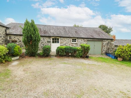 A stone building with a gray roof a white door green garage doors and bushes in front at Nant Bach Boduan near Nefyn