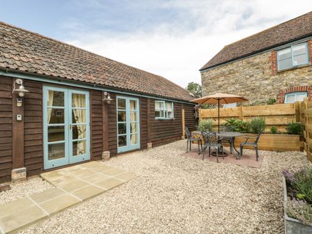 An outdoor patio area with gravel ground a table with umbrella and chairs next to wooden buildings at Toms Barn in Mudford Sock near Yeovil Somerset