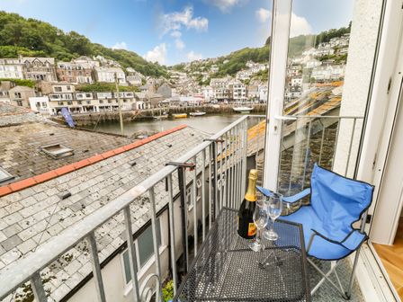 A small balcony with a black metal table holding a bottle of champagne and two glasses and a blue folding chair overlooking rooftops and a harbor with houses in the background at Harbour Hideaway in Looe