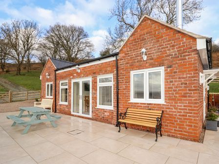 An outdoor patio area with a brick building a wooden bench a picnic table and wooden chairs at Border View Lodge in Llandrinio
