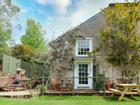 A stone cottage exterior with a white door and window a patio with wooden chairs and tables and green fencing surrounded by plants and trees at Wisteria Cottage in Marldon