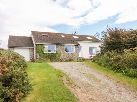 A house with stone and white exterior walls fronted by a gravel driveway and surrounded by bushes and grass at Cerrig in Rhoscolyn