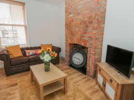 A living room with a brown sofa pillows flowers on a table a brick fireplace with a clock and a television on a wooden cabinet at Laridae in Whitby