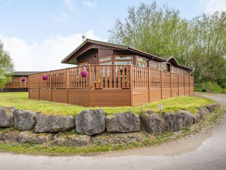 A wooden lodge with a fenced porch on a grassy area with large rocks and trees at Lakeview Lodge in South Lakeland Leisure Village