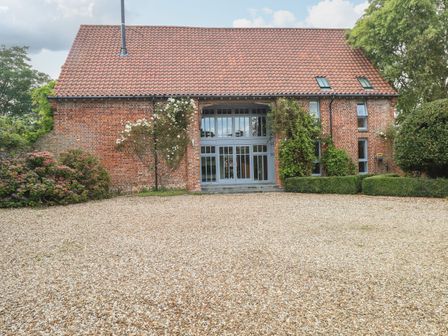 An exterior view of a brick house with a tiled roof and large windows with climbing plants on the walls and a gravel driveway at The Barn in Tunstead near Hoveton & Wroxham