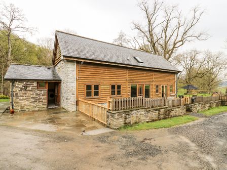 An exterior view of a barn style house with wooden siding stone walls fenced patio and surrounding trees at Ploony Barn in Knighton