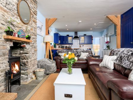 A living room with a stone fireplace a lit wood stove two brown leather sofas a white coffee table with flowers and a kitchen with blue cabinets at Sealark in Salcombe