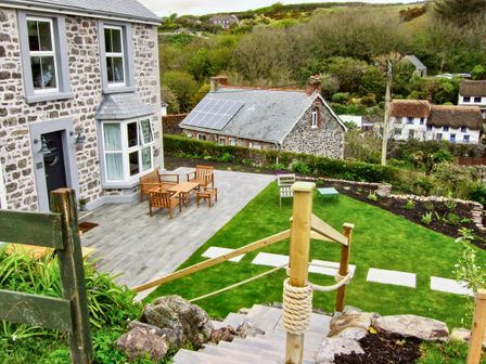 A stone house with wooden outdoor furniture on a patio and a garden with a stone path at Louvain in Cadgwith near Ruan Minor