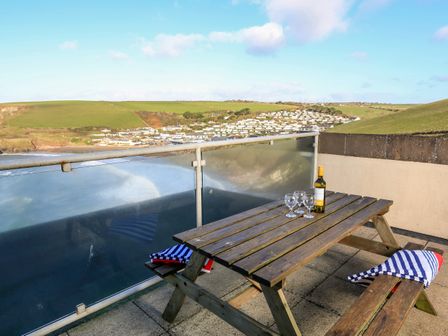 A wooden picnic table with a bottle of wine and four glasses on a balcony overlooking a coastal village and green hills at 2 The Bay in Bigbury