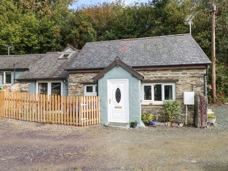 A stone cottage with a white door and wooden fence surrounded by trees at Pendre Cottage in Cenarth