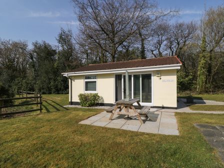 A small single-story house with a patio table and umbrella on a grassy lawn surrounded by trees at Ballasalla in Liskeard